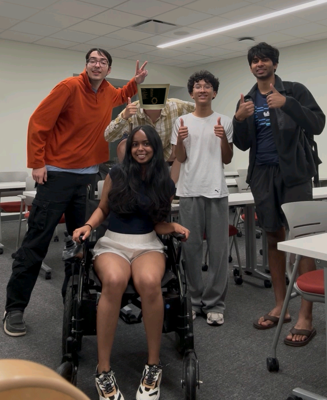 Five NeuroTech@VT members posing together in the lab classroom, one seated in the wheelchair prototype.